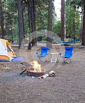 Campsite in the woods with a tent,two chairs, hammock and a burning firepit.