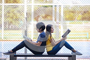 On a campus, a couple of students are studying together, and a teenager sits on a seat beside a sports court with a book