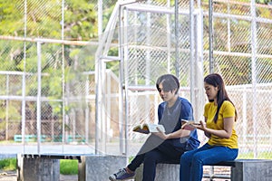 On a campus, a couple of students are studying together, and a teenager sits on a seat beside a sports court with a book