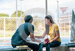 On a campus, a couple of students are studying together, and a teenager sits on a seat beside a sports court with a book