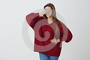 Can I look now. Curious and excited attractive woman in trendy loose red sweater covering eyes with hand and standing