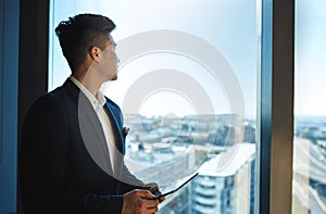 He can see success on the horizon. a young businessman using a digital tablet while looking out the window in an office.