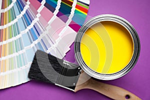 Can with yellow paint, color palette and brush on purple background, flat lay