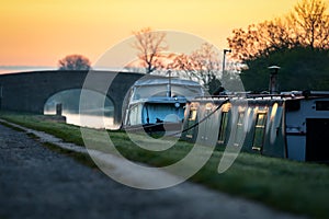 Canal boat and river vessel moored overnight early morning sunrise standing on tow path with golden sunlight in sky silhouettes