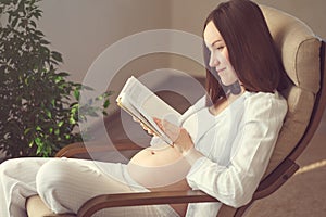 Candid cozy portrait of a pregnant woman resting at home and reading a book on a rocking chair by the window