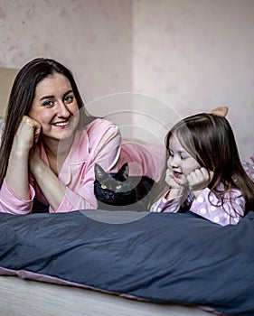 Candid family spending time together. Beautiful mother in pajamas, little daughter and black cat in bed. Happy family