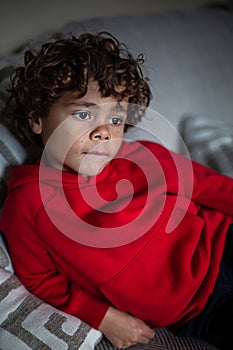 Candid photo of a young diverse African American little boy sitting on a couch at home watching streaming content