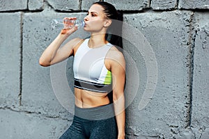 Candid portrait of fitness young woman drinking water from the bottle after training outdoors. Athlete female taking a break after