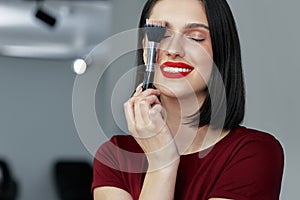 Candid portrait of a gorgeous brunette female with red lips posing with a brush after making make-up tutorial. Happy young woman
