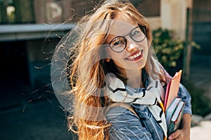 Candid portrait of a happy young student woman with long red hair smiling and wearing transparent eyeglasses standing next to the