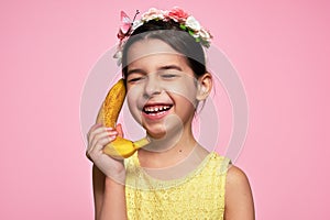 A candid portrait of a joyful smiling little girl holds a banana, like a phone, isolated on pink background. A happy kid in a