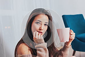 Candid portrait of sensual smiling asian girl young woman with dark long hair in cozy beige sweater holding cup of tea on white
