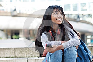 Young Indian woman sitting on steps with mobile phone and looking away