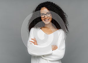 Candid shot of beautiful African American woman with curls, has gentle smile, keeps hands crossed, wears white soft sweater,