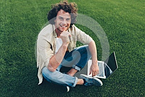 Candid shot of cheerful freelance man with curly hair using laptop for working outdoors, connected to free wireless, sitting on