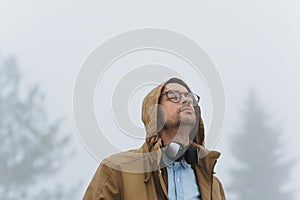 Candid shot of smart male listening to music using his headphones and mobile phone, looking up and wearing hood of coat and