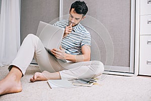 Candid shot of young Caucasian man student sitting on the floor in the room in front of open laptop computer. Serious European