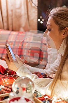 Candid young smiling woman communicates with friends using a smartphone texting message at home in a cozy bed on a red checkered b