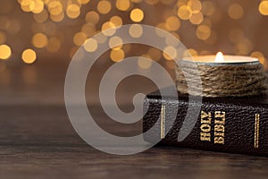 Candle burning and closed holy bible book on wooden table with bokeh background