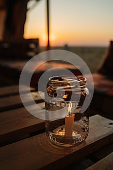 Candle burning inside a glass jar on a table. Sun setting over a steppe horison