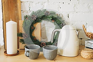 Candle, christmas wreath of fir branches, teapot and cups on kitchen, cozy home scandinavian design