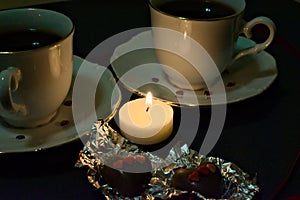 A candle illuminates two cups and two chocolate hearts. Valentine`s Day.