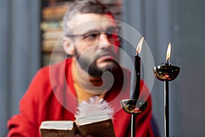 Bearded diviner sitting near burning candles and reading ancient book