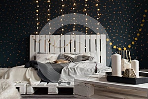 Candles on a table focus. Cosy bedroom interior with a white wooden bed, plaids, flashlights, candles and dark walls. Christmas de