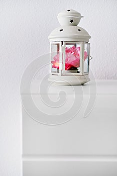 Candlestick lantern shape decorated with pink artificial flower stands on bedside table or nightstand against white color wall