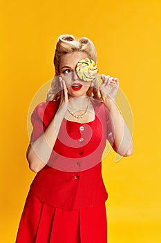 Candy lover. Portrait of beautiful young blonde girl with stylish hairstyle in red suit posing against yellow studio