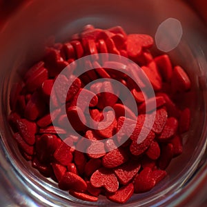 Candy sprinkles in shape of red hearts in glass on background.
