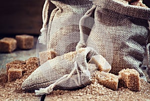 Cane brown sugar cubes and crystals in burlap sacks on old wooden background in rustic style, selective focus