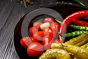 Canned cherry tomatoes, cucumbers, peppers in a round black plate on a dark background, close-up