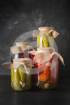 Canned fruits and vegetables, plum, cucumber, tomato in glass jars on black wooden board.