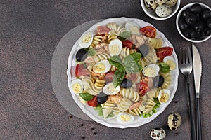 Canned salmon salad with pasta, cherry tomatoes, quail eggs, basil and black olives on brown background, Top view