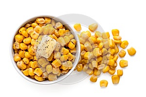 Canned sweet corn with a knob of butter and ground black pepper in a white ceramic bowl next to spilled sweet corn isolated on