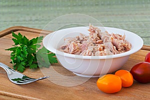 Canned tuna fillet in white porcelain bowl on a brown wooden cutting board with some cherry tomatoes, parsley and fork. Seafood,