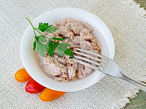 Canned tuna fillet in white porcelain bowl, fork, parsley and some cherry tomatoes on a beige table table napkin. Seafood, healthy