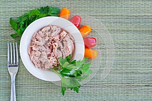 Canned tuna fillet in white porcelain bowl, fork, parsley and some cherry tomatoes on a green table mat made of natural plant