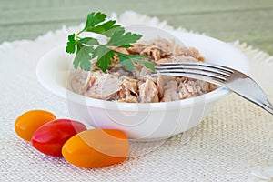 Canned tuna fillet in white porcelain bowl, fork, parsley and three cherry tomatoes on a beige table table napkin. Seafood,