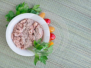 Canned tuna fillet in white porcelain bowl, parsley and some cherry tomatoes on a green table mat made of natural plant fibers.