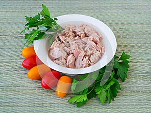 Canned tuna fillet in white porcelain bowl, parsley and some cherry tomatoes on a green table mat made of natural plant fibers.