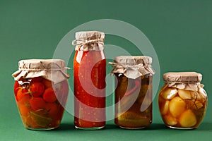 Canned vegetables in jars on a green background. Pickled cucumbers, tomatoes, garlic, chili sauce