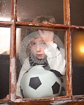 Cant it rain some other time. a sad little boy holding a soccer ball while watching the rain through a window.