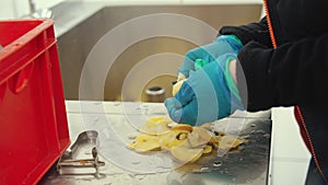 Canteen worker in blue gloves peeling potatoes with a knife and reaching for another potato from a washer. Dining hall