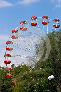 Cantilevered observation wheel with empty red cabins on blue sky and green trees in park photo