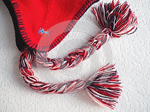 Cap with earflaps in the Norwegian style. Knitted fashion item with ties and pom-pom tassels. Red yarn with black and