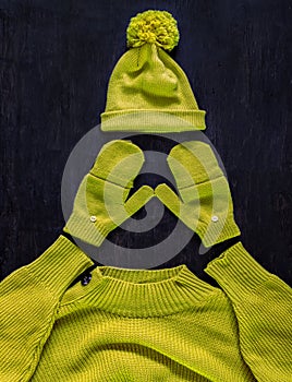 Cap with pompon, mittens, sweater in yellow-green color on a dark wooden background. Top view, flat lay.Concept stylish youth warm