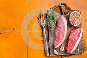 Cap Rump steak, raw beef meat steak with thyme and salt on wooden butcher board. Orange background. Top view. Copy space