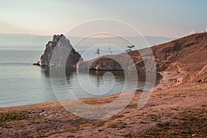 Cape Shamanka rock in clear blue Lake Baikal among grassy steppe, mountains, warm sunset